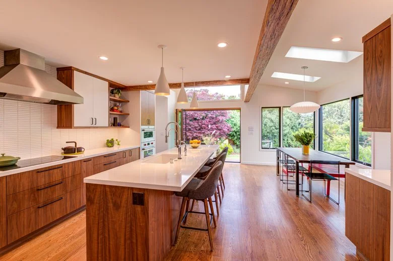 Open kitchen and dining area with walnut cabinetry, a large island with seating, skylights, and clerestory windows that bring daylight deep into the expanded main level.