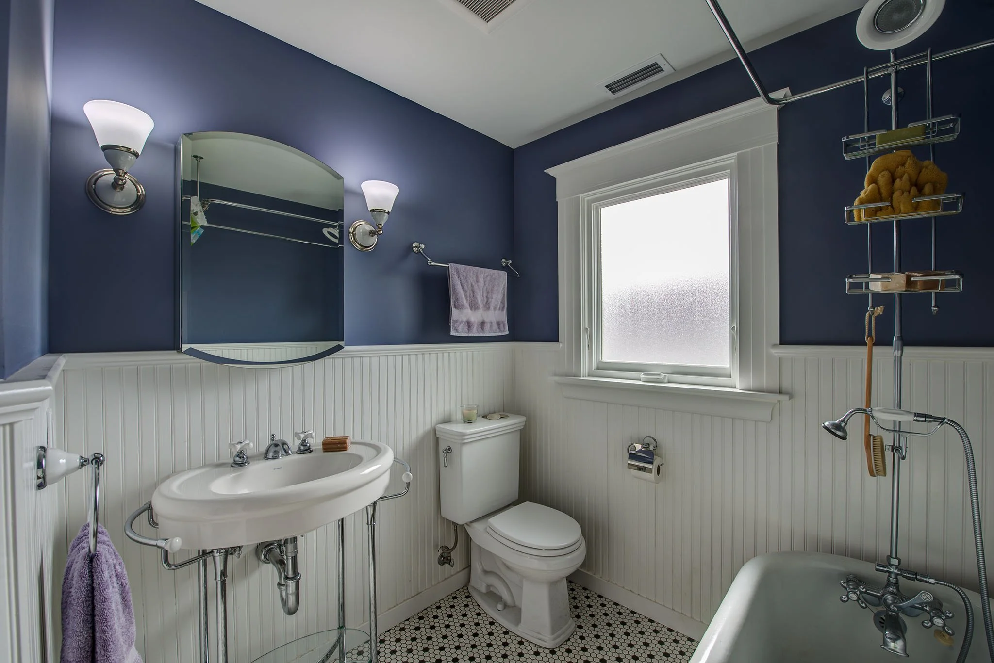 New bathroom with deep blue walls, beadboard wainscoting, pedestal sink, hex tile floor, and a tub with exposed shower by a frosted window for soft light.
