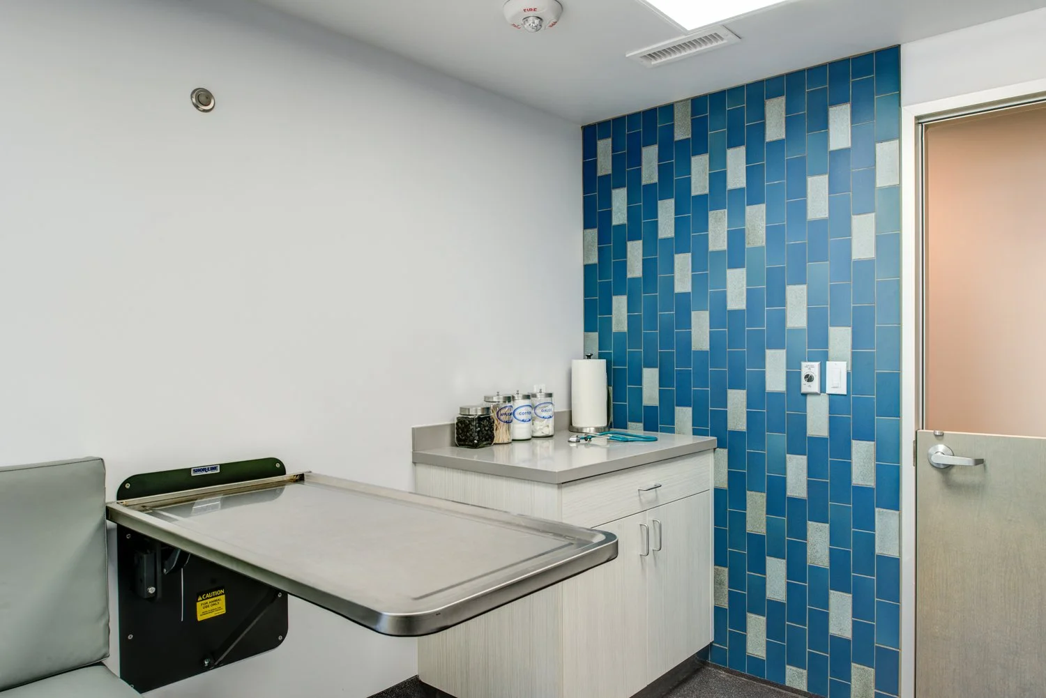 Remodeled exam room featuring a stainless exam table, light wood cabinetry, and a blue and gray tiled wall, designed for efficient workflows and easy sanitation.