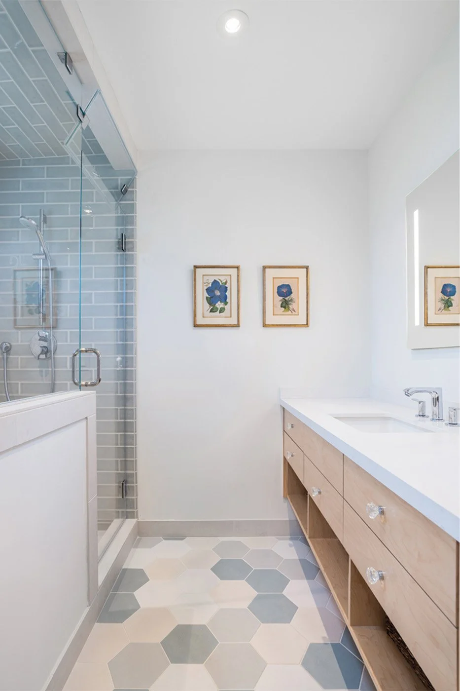 Contemporary bathroom featuring a glass-enclosed shower with blue-gray tile, light wood vanity with white countertop, and patterned hexagonal floor tiles in neutral tones