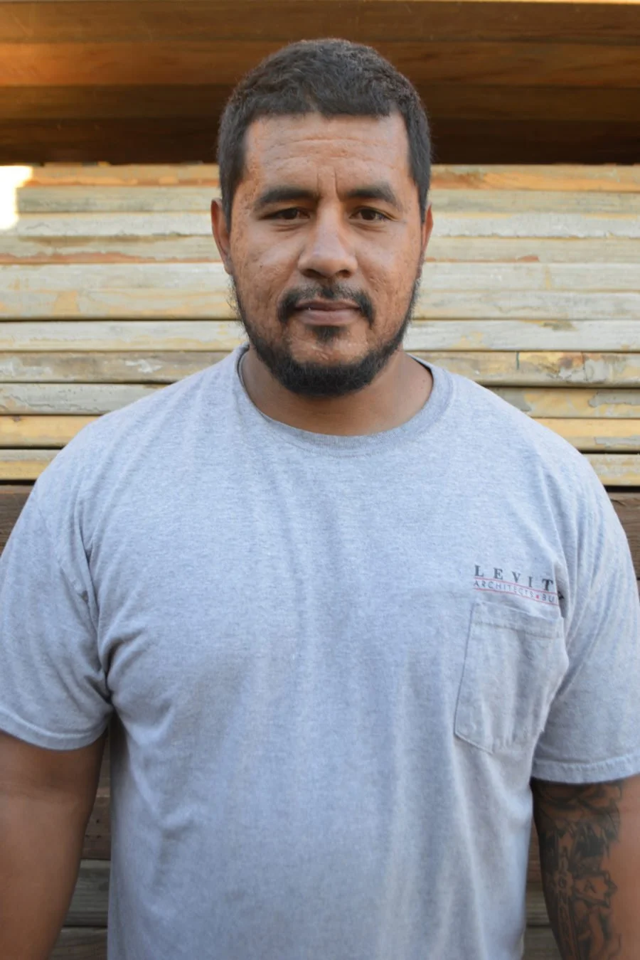 A man with dark hair and a beard wearing a light gray t-shirt standing in front of a wooden and stone background.