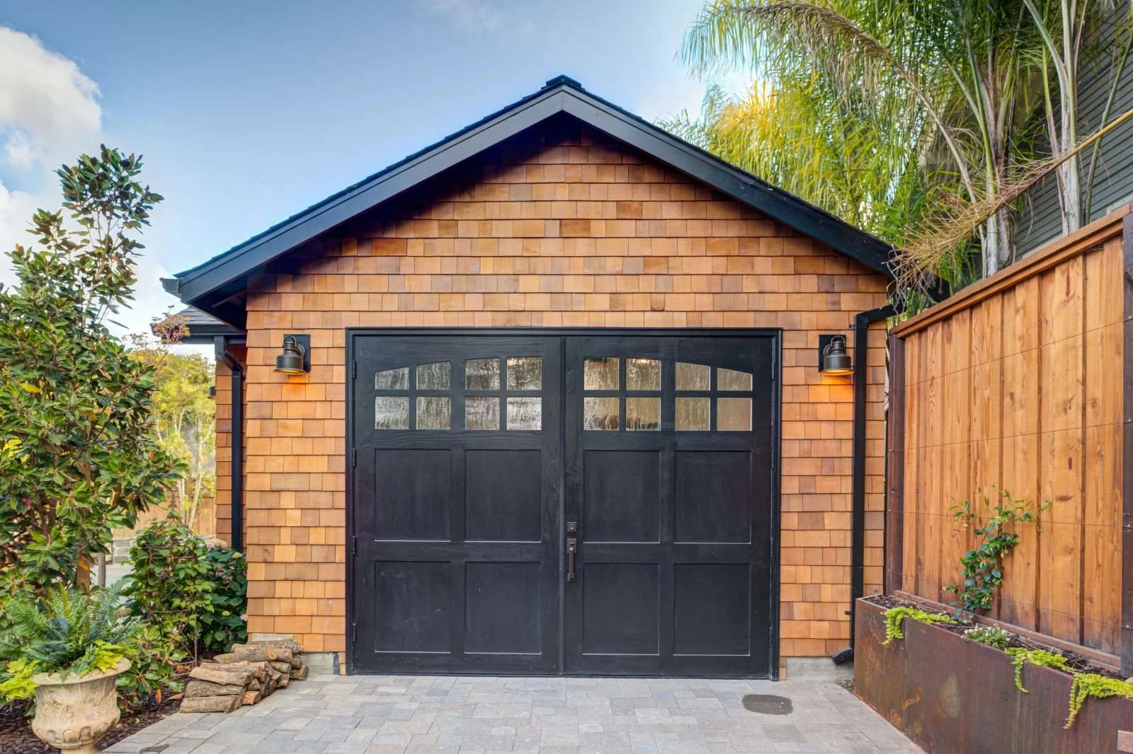The accessory structure’s exterior is a compact, gabled volume clad in cedar shingles with dark-painted wood doors, reinforcing a warm material palette while serving as a flexible studio or "Man Cave".