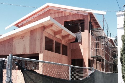 Front exterior during construction with plywood sheathing, new roof framing, and scaffolding, showing the house rebuilt to the floor structure for the second story addition.