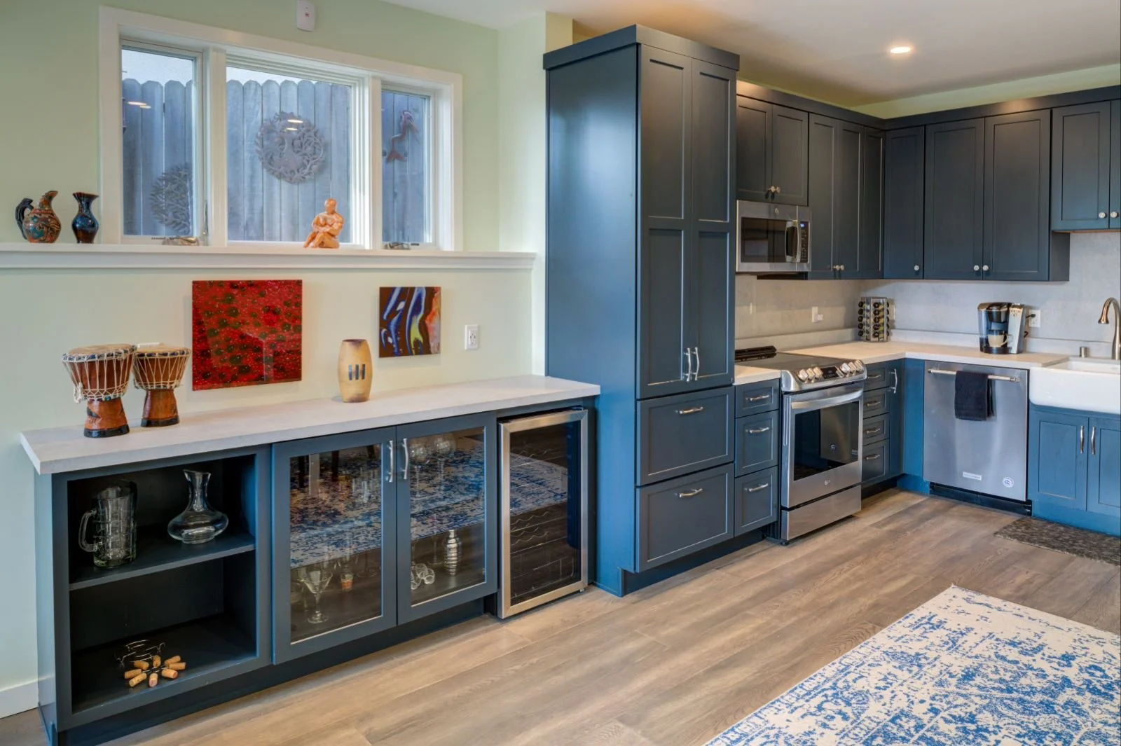 Open kitchen and bar area in Levitch’s Grand Lake ADU featuring blue cabinetry, quartz counters, wine fridge, display shelving, and art beneath a wide window.