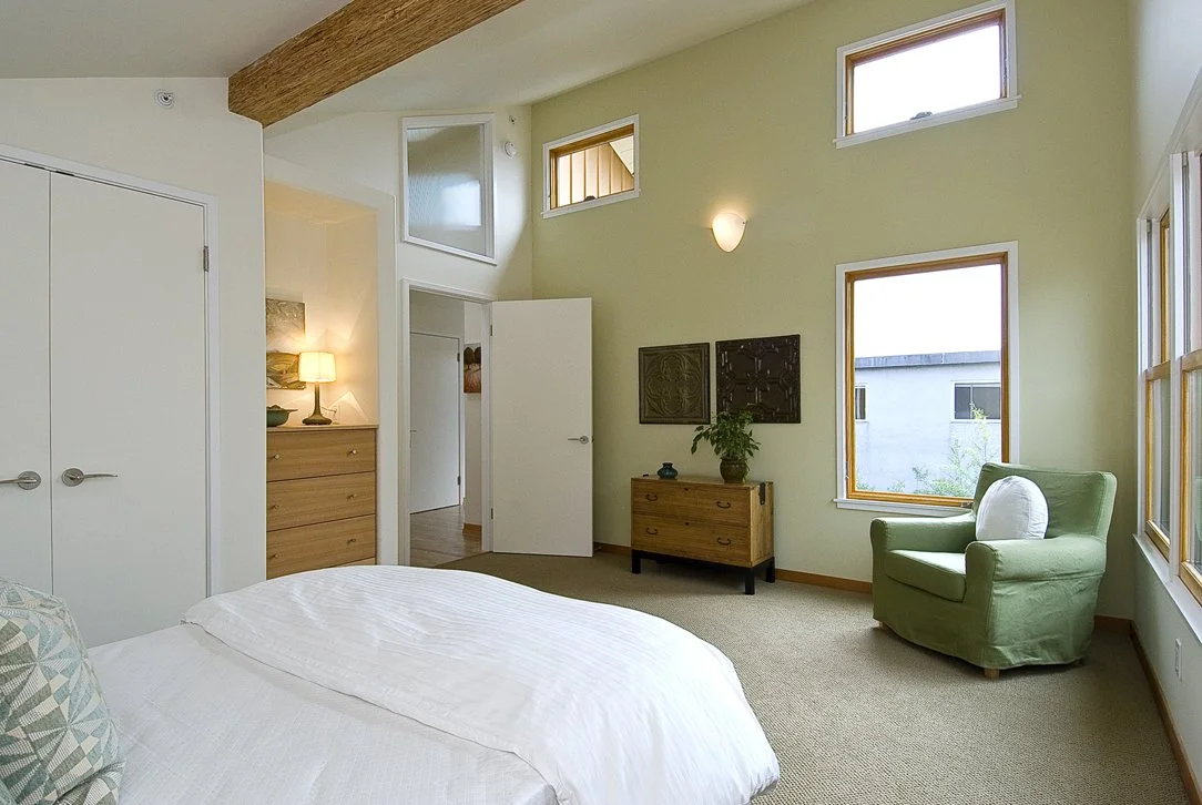 Primary bedroom with vaulted ceiling, exposed wood beam, soft green walls, and multiple windows that provide natural light and ventilation in this energy-conscious new construction.