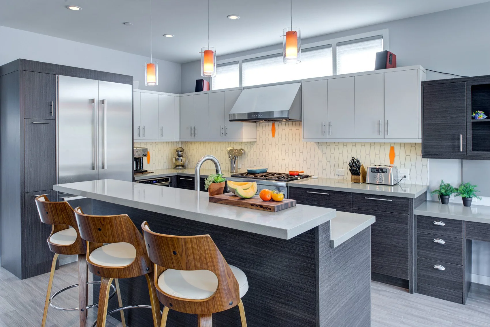 Warm modern kitchen with a large quartz island and seating, two-tone cabinetry, stainless range and hood, and geometric tile backsplash lit by simple pendant lights.