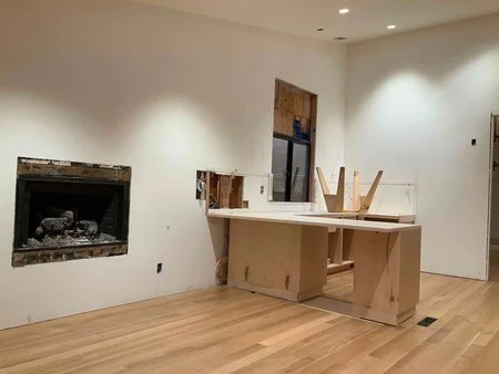 Family room during construction with new wood floors installed, fireplace opening exposed, and kitchen peninsula framing underway as part of the interior remodel.