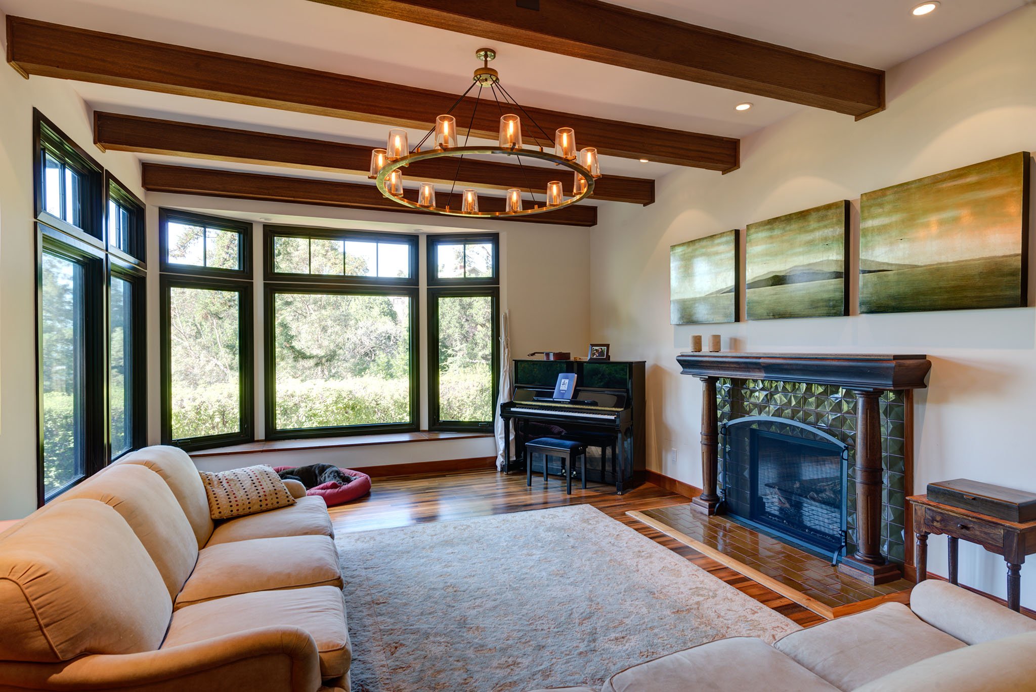 Bright living room remodel with exposed wood ceiling beams, large woodland windows, and a craftsman style fireplace surround, creating a warm space for family gathering.