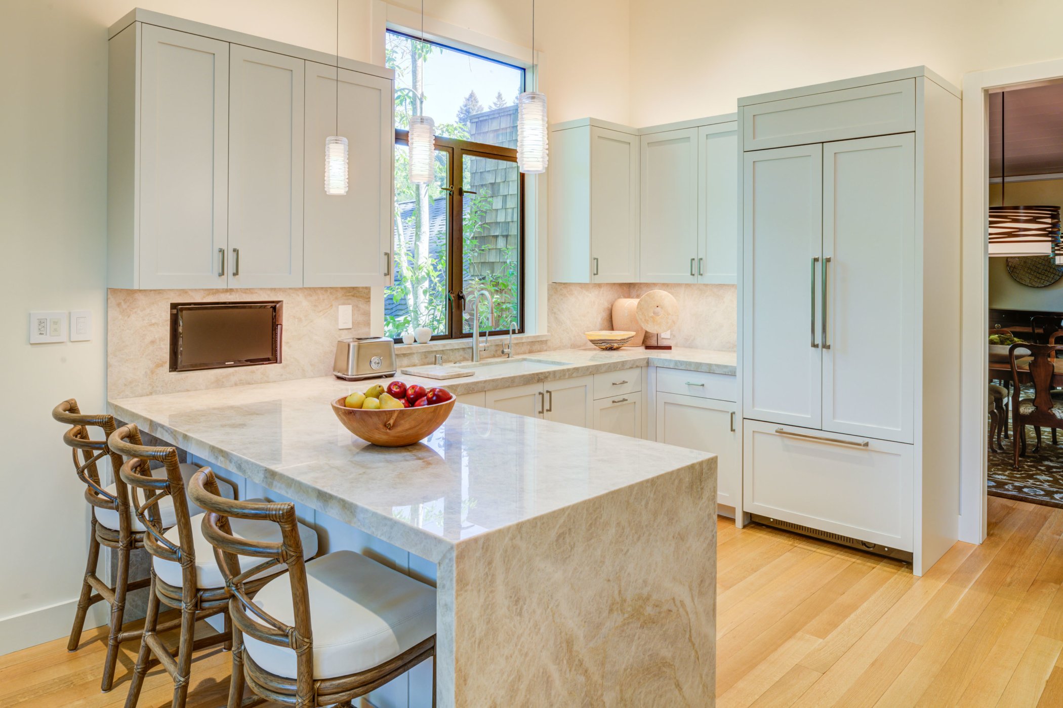 Bright remodeled kitchen with pale custom cabinetry by Levitch, a stone peninsula with seating, pendant lights, and a larger window over the sink, bringing in natural light.