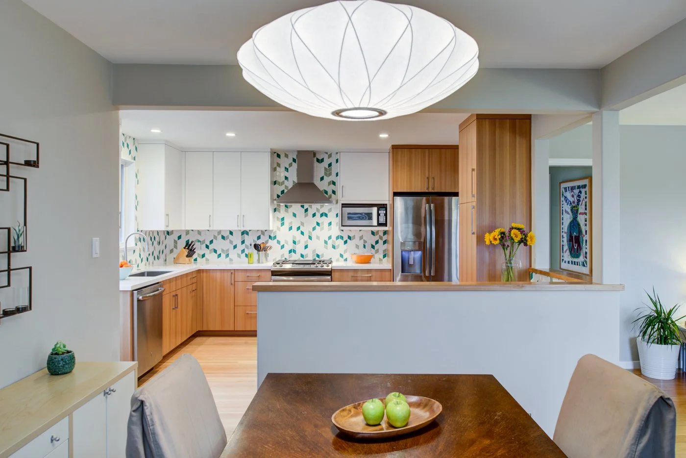 View from the dining table toward the open kitchen, showing modern pendant light, Levitch built eucalyptus cabinets, stainless appliances, and green patterned tile backsplash.