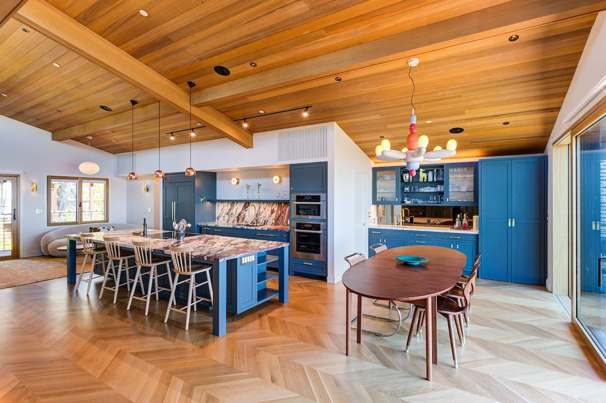 Rich blue cabinetry, dramatic veined stone, warm oak flooring, and a wood-clad ceiling establish a layered, contemporary aesthetic. Exposed beams and the open-plan layout reinforce the Kitchen as the social center of the home.