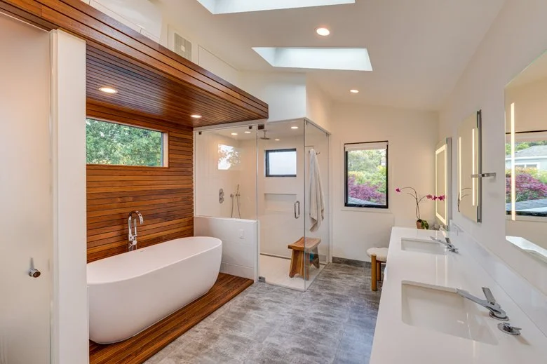 Primary bathroom featuring a freestanding soaking tub, wood accent wall, glass shower enclosure with bench, and skylights that create a calm and spa-like retreat.
