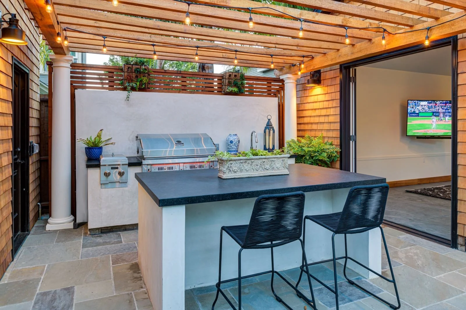 Outdoor kitchen covered by a wood pergola, stone pavers, and a built-in island clad in smooth plaster. Durable countertops, bar seating, and an integrated grill support casual dining and social gathering.