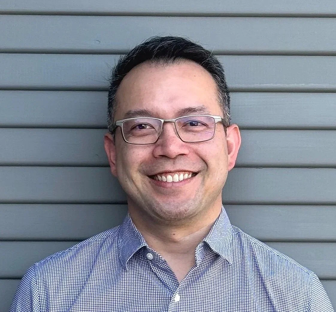 A smiling man with glasses wearing a blue and white checkered shirt standing in front of a gray horizontal panel wall.