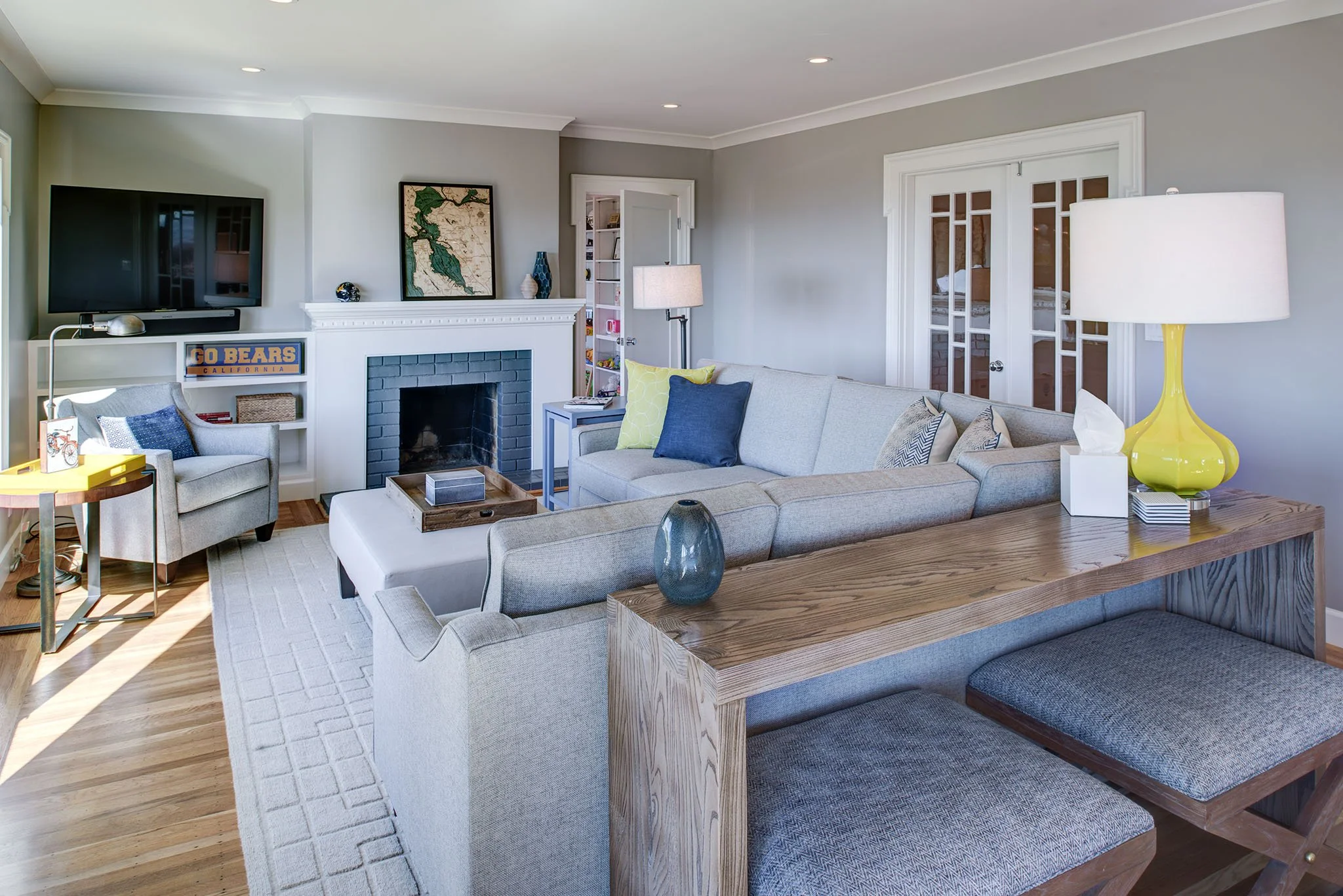 Remodeled living room with a painted brick fireplace, built in media storage, and a sectional sofa with blue and gold accents. Neutral finishes keep the space cohesive with the open plan layout.