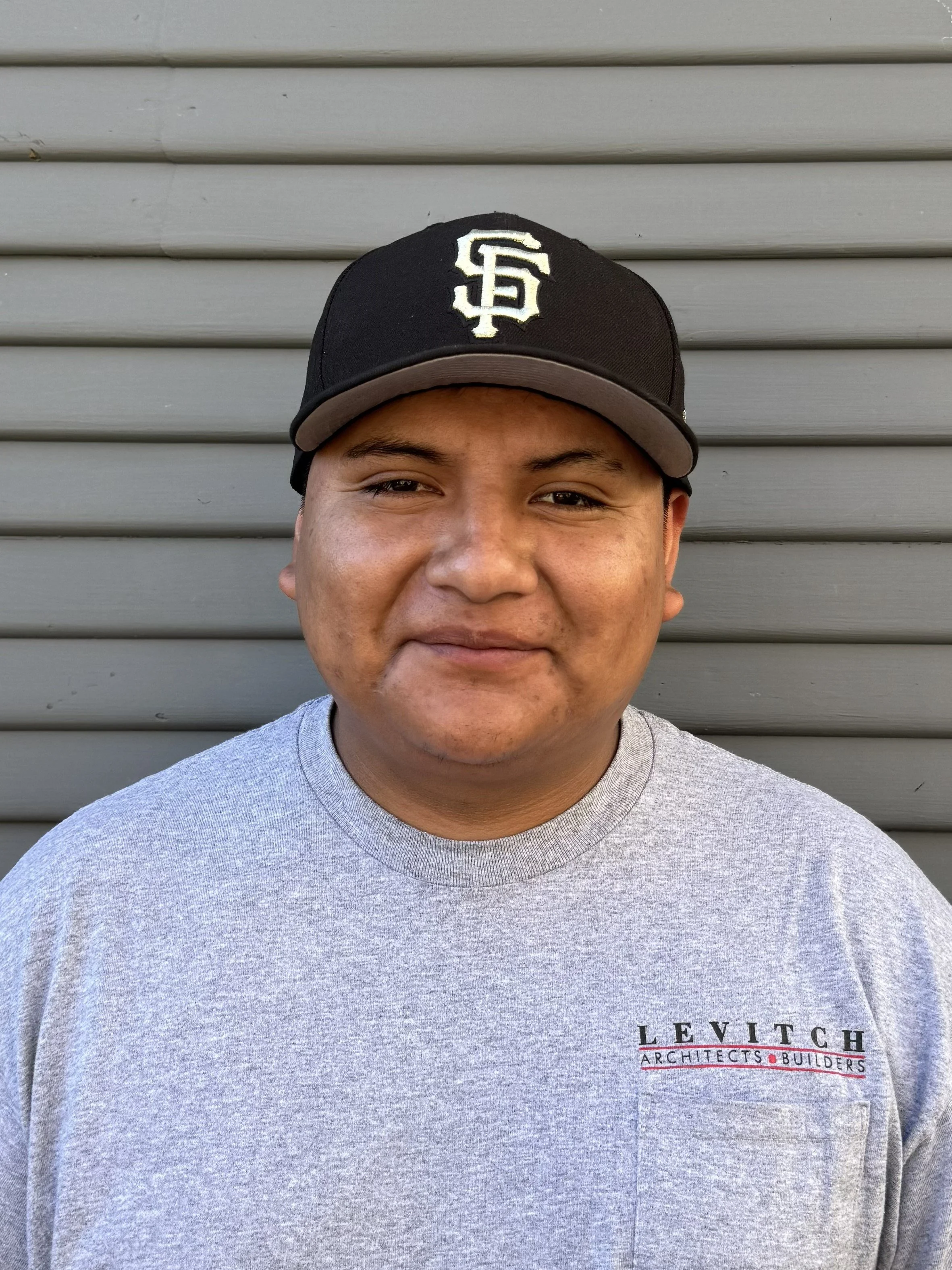 A young man wearing a black baseball cap with white embroidered letters 'SF' standing in front of a gray slatted wall, smiling slightly, wearing a gray T-shirt with 'LEVI TCH ARCHITECTS BUILDERS' printed on it.