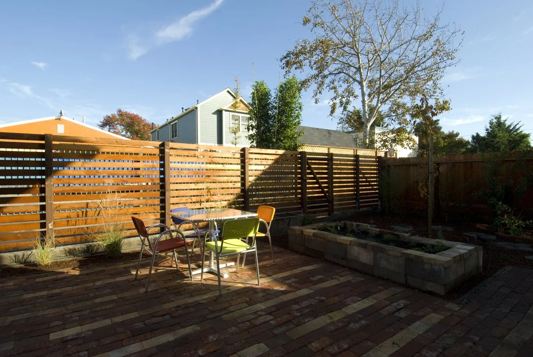 Rear yard with paved patio, planting beds, and wood privacy fencing, designed as a low maintenance outdoor space that complements the sustainable home design.