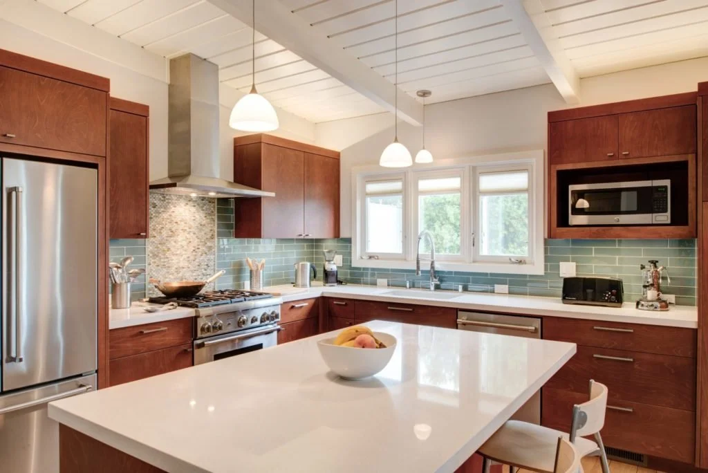 Wide view of the remodeled kitchen with a large white island, blue-green tile backsplash, slab front cabinetry, new windows, and a sloped wood ceiling that keeps the midcentury character.