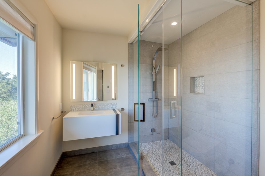 Modern bathroom with floating vanity, illuminated mirror, and glass-enclosed shower, lit by a window and skylight.