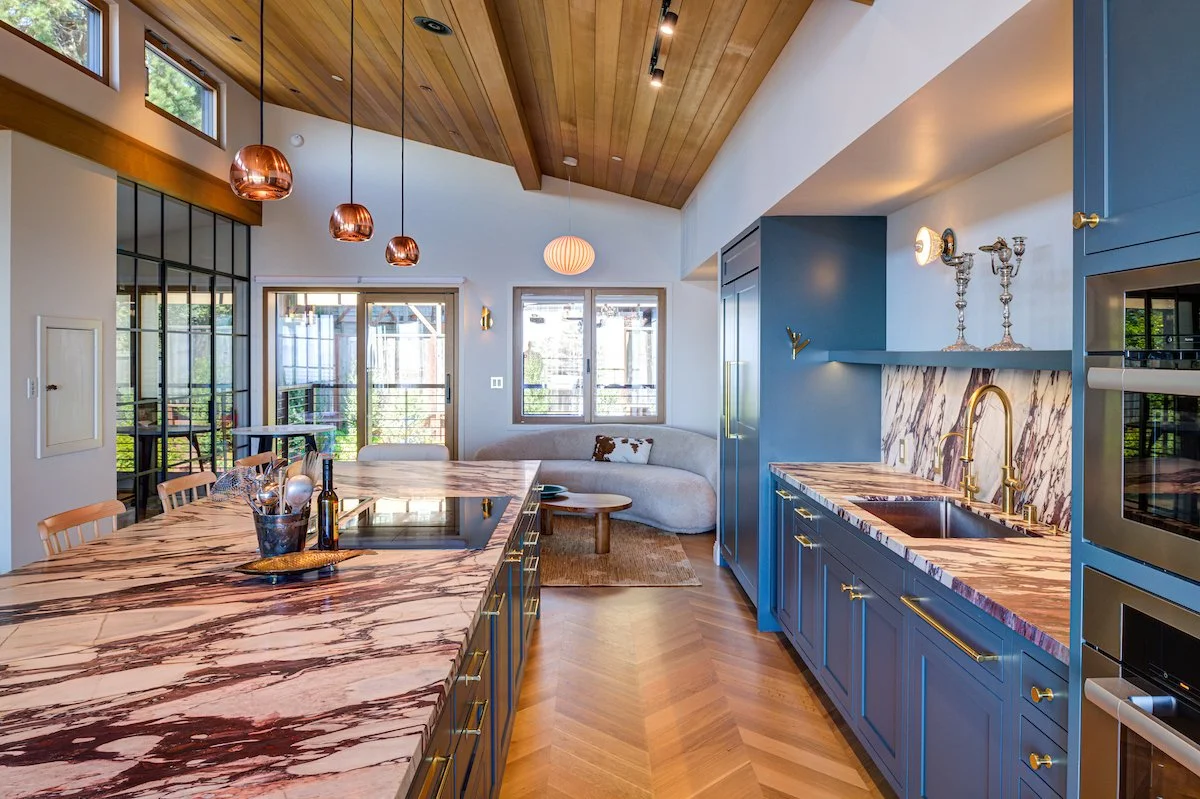 Custom blue cabinetry is paired with highly expressive stone countertops and backsplash, creating a bold focal point against the warm oak floor and ceiling. Integrated appliances and custom Levitch made cabinetry maintain a clean, cohesive compositio