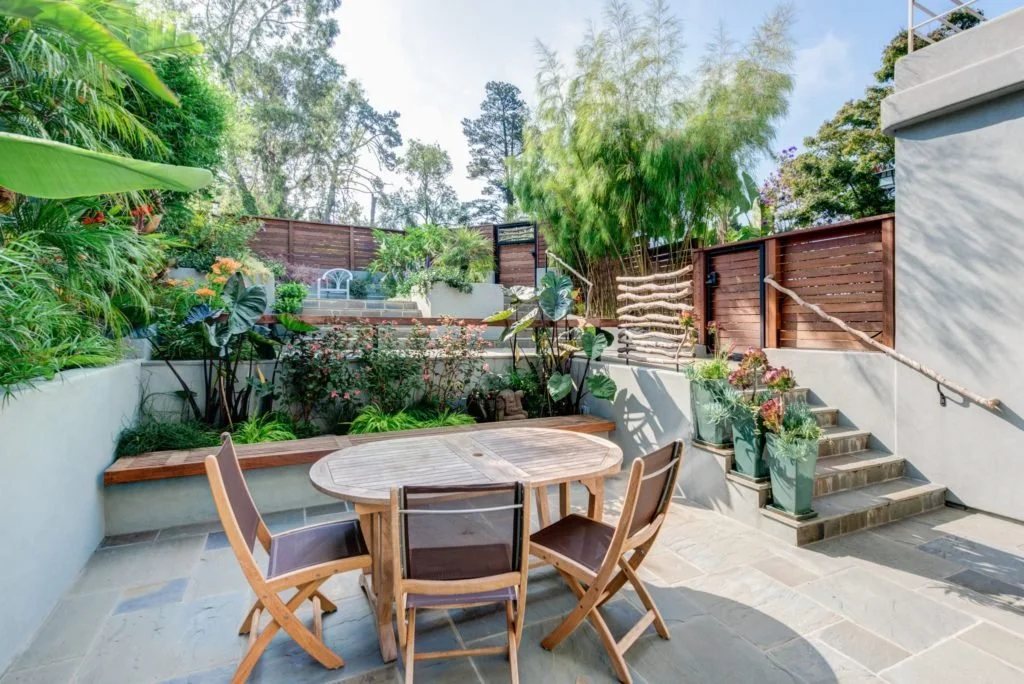 Lower terrace patio with a round dining table, built in planter seating, stone paving, and stairs with branch inspired handrails, creating outdoor entertaining space on a sloped yard.