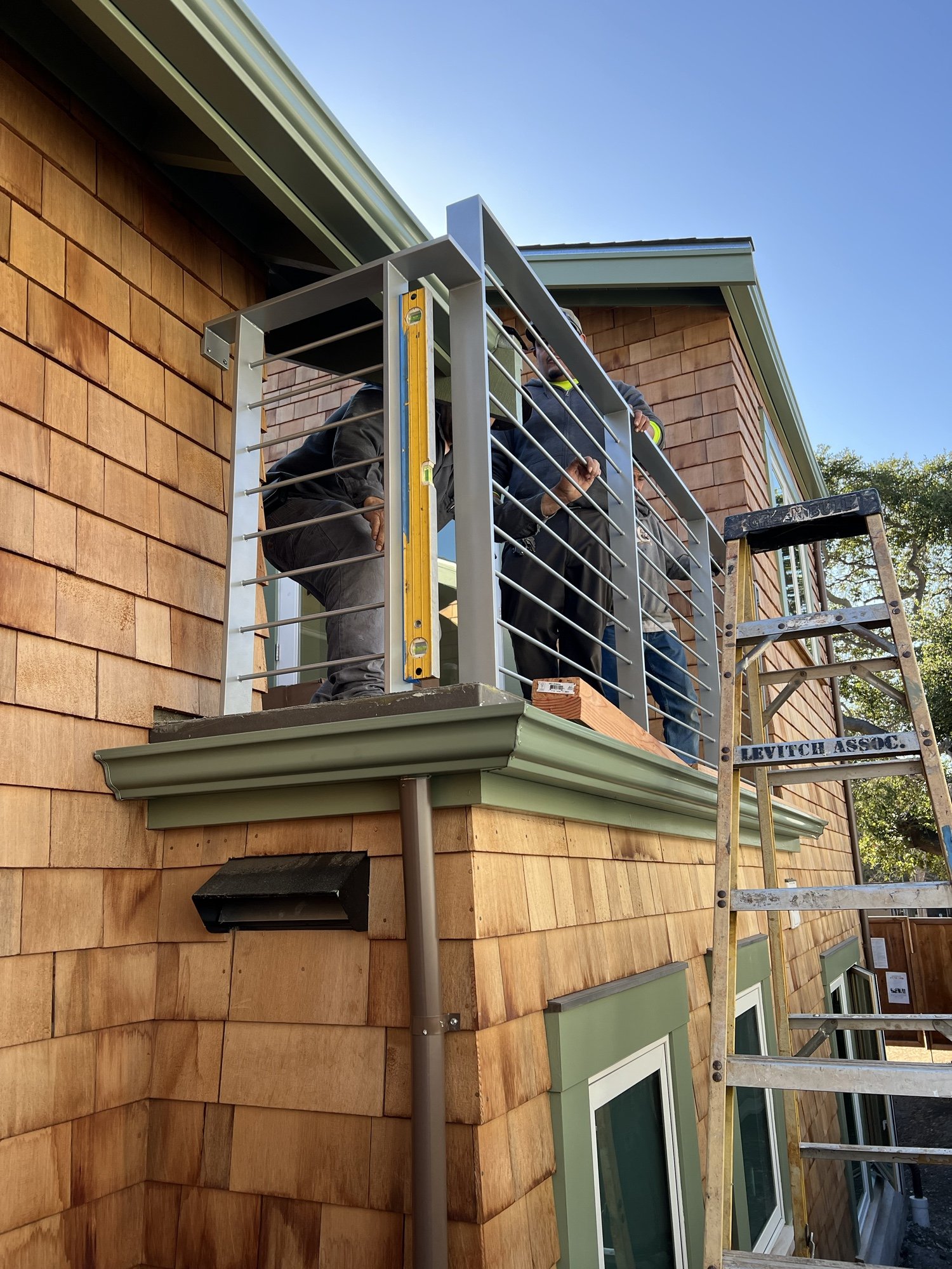 Exterior view of the new upper level deck under construction, with workers installing a modern metal railing. The deck connects to a new bedroom and expands outdoor living on the compact urban lot.