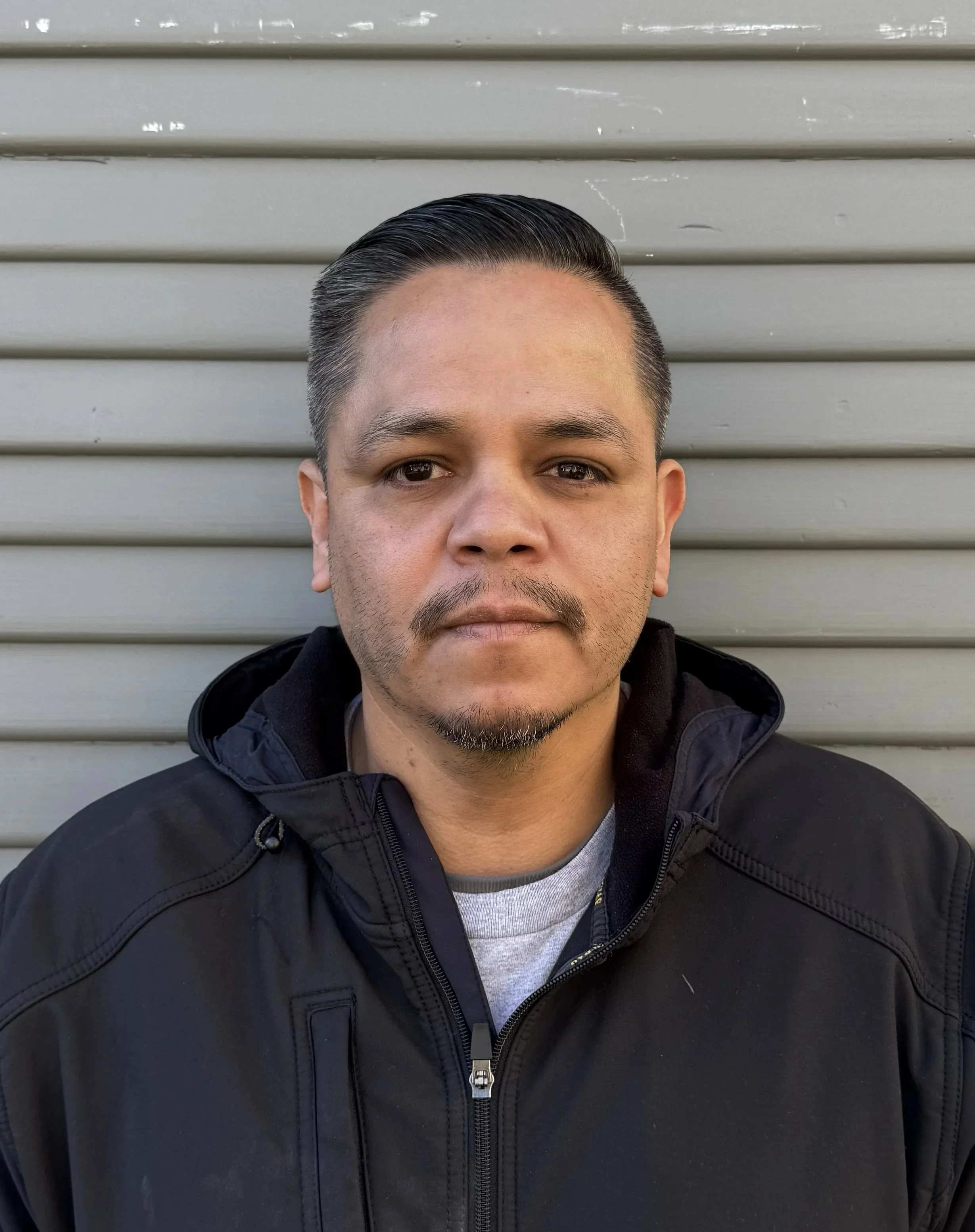 A man with short, dark hair and a goatee, wearing a black jacket, standing in front of a gray, horizontal metal siding background.