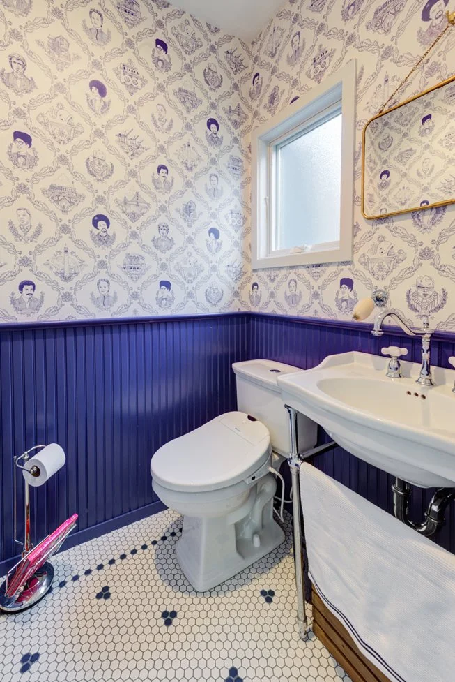 Small powder room with playful illustrated wallpaper, deep purple beadboard wainscoting, classic hex tile floor, pedestal sink, and a frosted window for soft natural light.