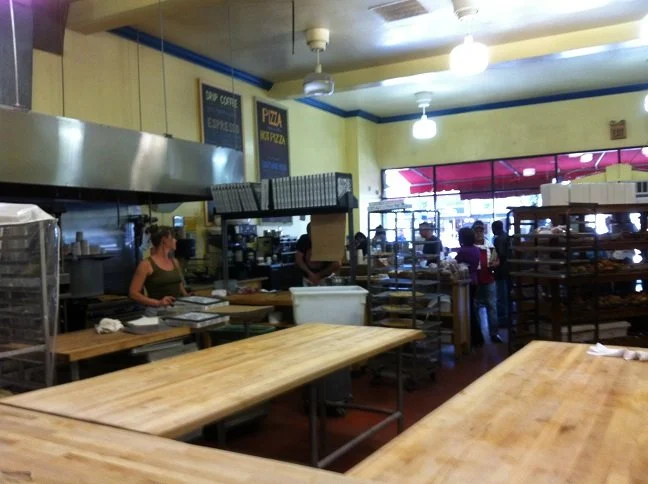 Open bakery production space with large wood prep tables, commercial ovens, and staff circulation paths, showing a functional layout designed for efficient bread and pastry preparation.