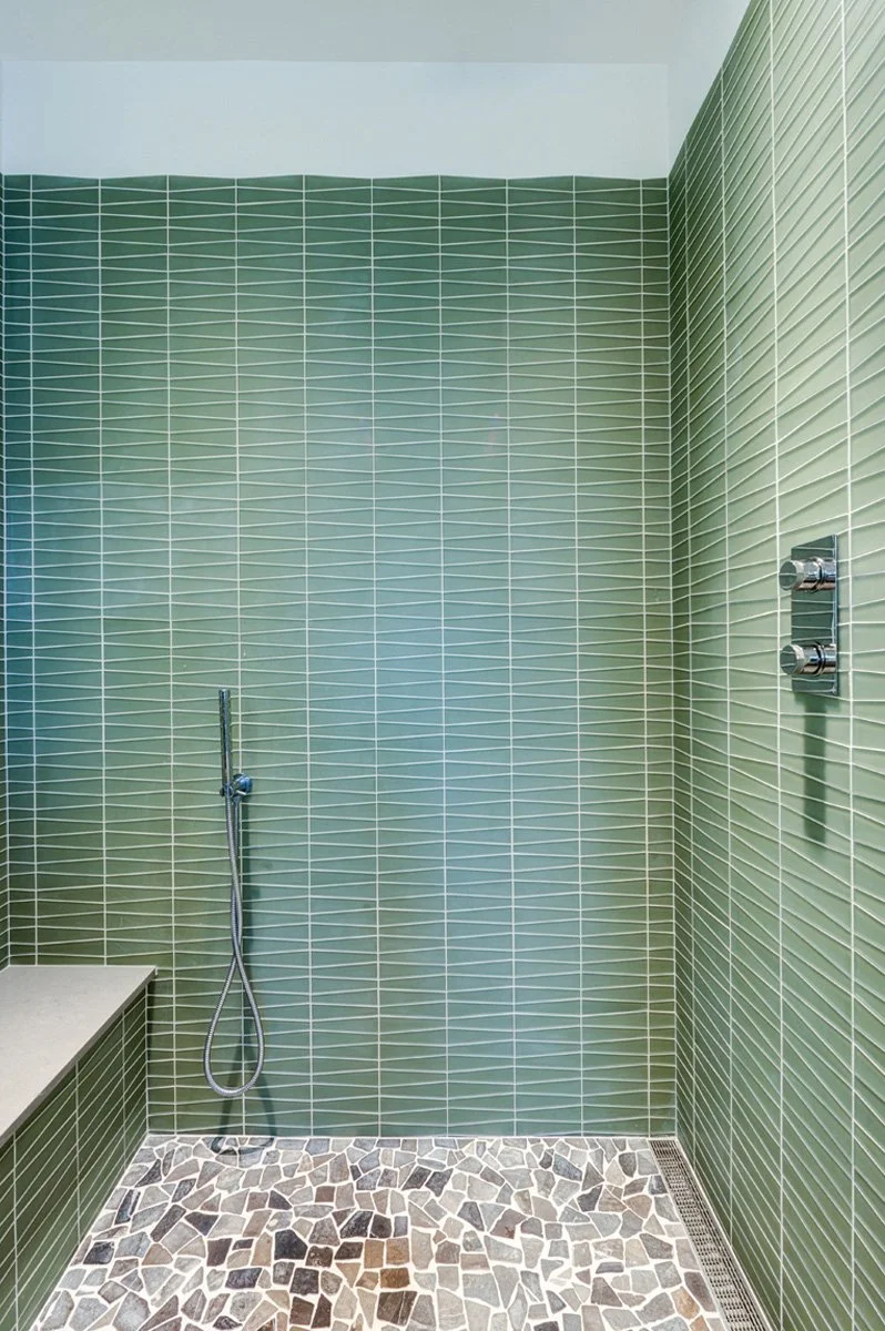 Close-up of the enlarged walk-in shower in the remodeled primary bathroom, featuring green linear tile, a built-in bench, and pebble-stone shower flooring.