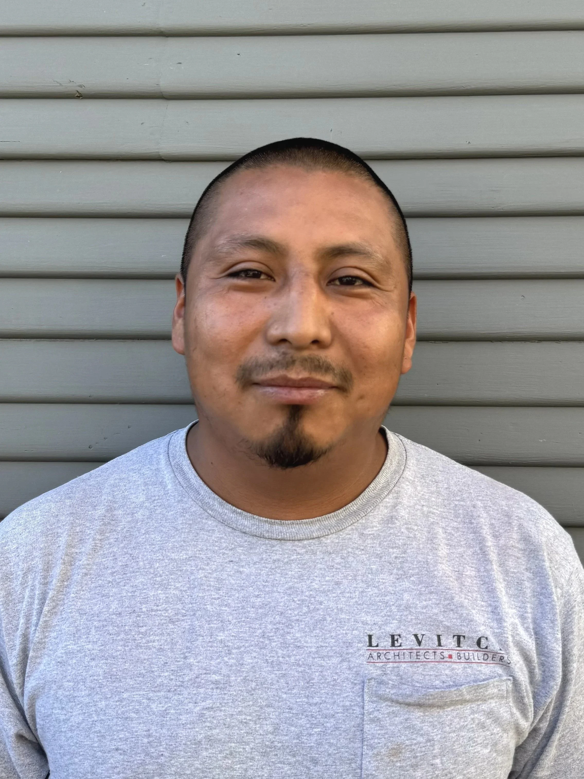 A man with short dark hair and a goatee smiling while standing outdoors in front of a light gray horizontal siding wall, wearing a gray T-shirt with the logo 'Levitc Architects + Builders' on the chest.