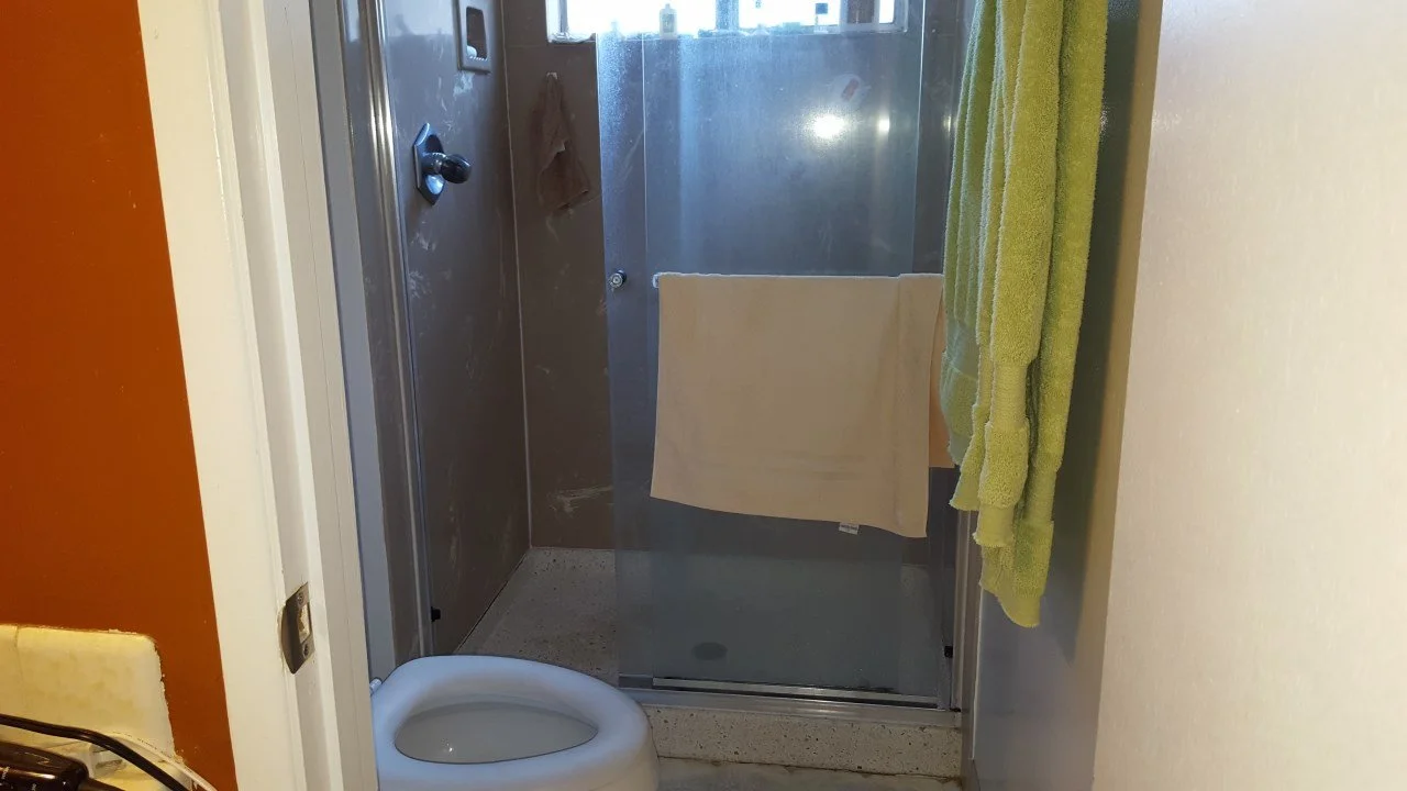 Existing primary bathroom showing a narrow shower stall with sliding glass door, dated fixtures, limited clearance, and low natural light prior to the Alameda waterfront remodel.
