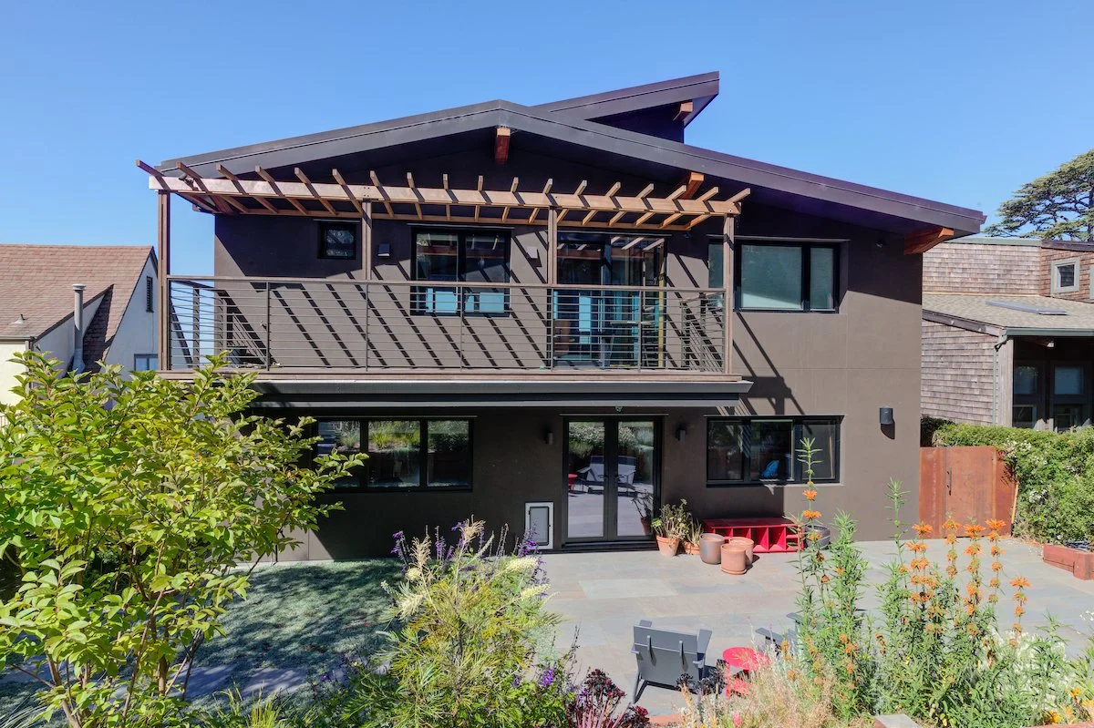 Rear view of the home with pergola-covered back deck and a patio covered in pavers to maximize usable space on this sloping lot.
