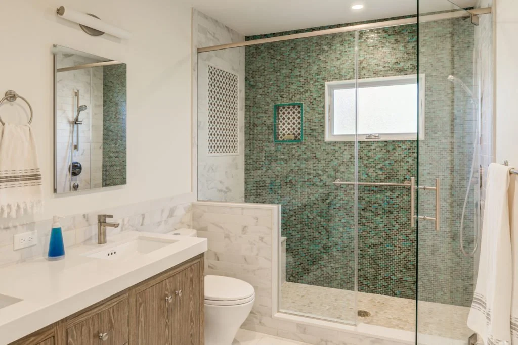 Remodeled bathroom with a wood vanity, clean white walls, and a glass enclosed shower finished in teal mosaic tile and marble look wainscot for a brighter, larger feel.