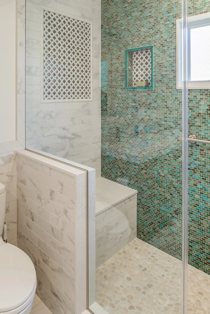 Bathroom shower detail with teal and blue mosaic tile, a recessed soap niche with patterned inset, marble look wall tile, glass enclosure, and pebble style shower floor.