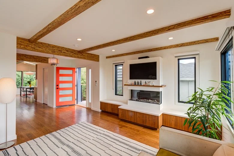 Living room view highlighting the relocated front entry with a bold orange door, custom media wall, and midcentury-inspired finishes that improve flow and natural light throughout the home.