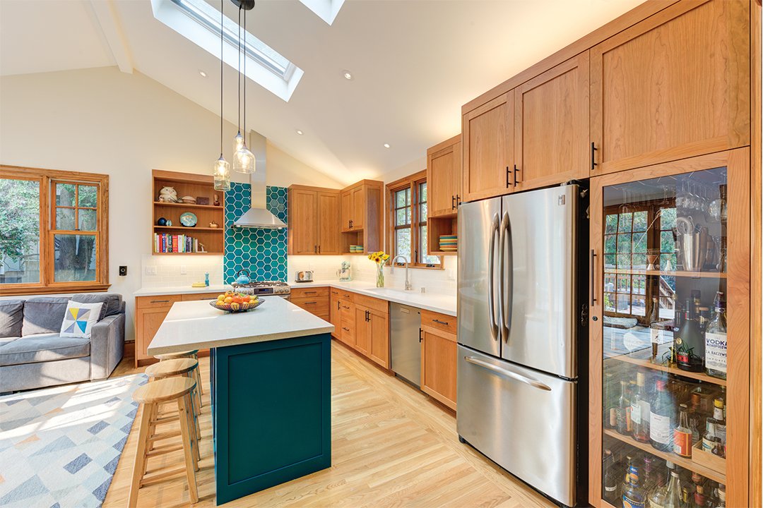 Bright remodeled kitchen with a central island painted turquoise, wood cabinetry, and stainless steel appliances, set beneath vaulted ceilings with skylights and large windows bringing in natural light.