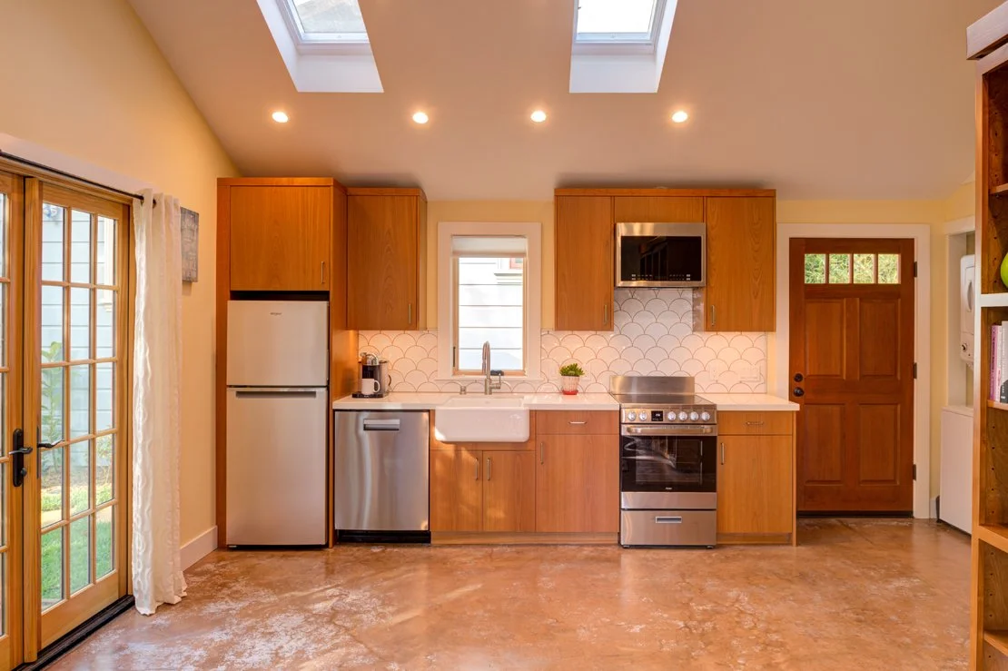 ADU Kitchen wall featuring tall custom cabinets in natural wood that extend to the top of the wall to maximize storage. Abundant natural light via windows, doors, and skylights. 
