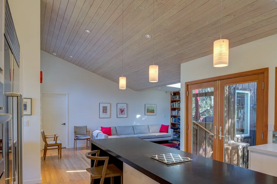 View from the kitchen island into the living area, showing vaulted wood ceilings, pendant lighting, and glass doors connecting the interior to the outdoors.