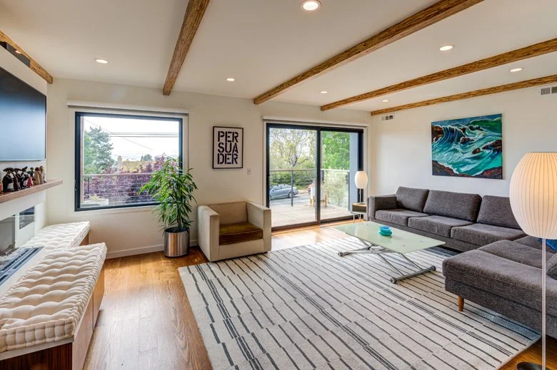 Living room interior with exposed wood ceiling beams, large windows, and sliding glass doors that open to the deck, creating a bright and comfortable gathering space connected to the outdoors.