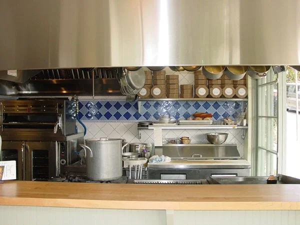 Open front kitchen at Grégoire Piedmont with a wood counter, stainless ventilation hood, and blue tile backsplash, set up for visible food prep and quick service at the window.