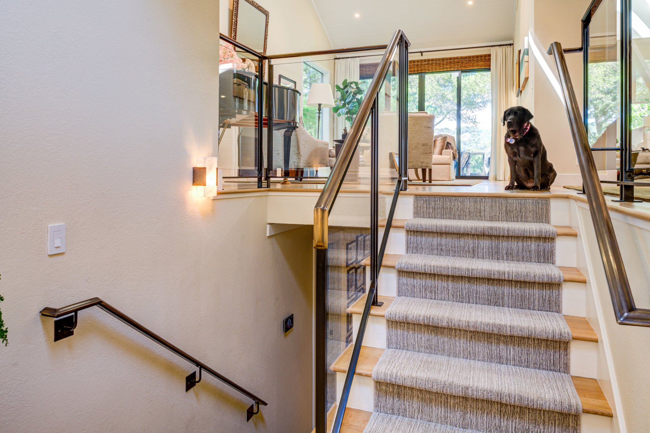 View up the interior stairs showing the custom glass and metal railing, carpeted steps, wall lighting, and the family room beyond with improved safety and openness.