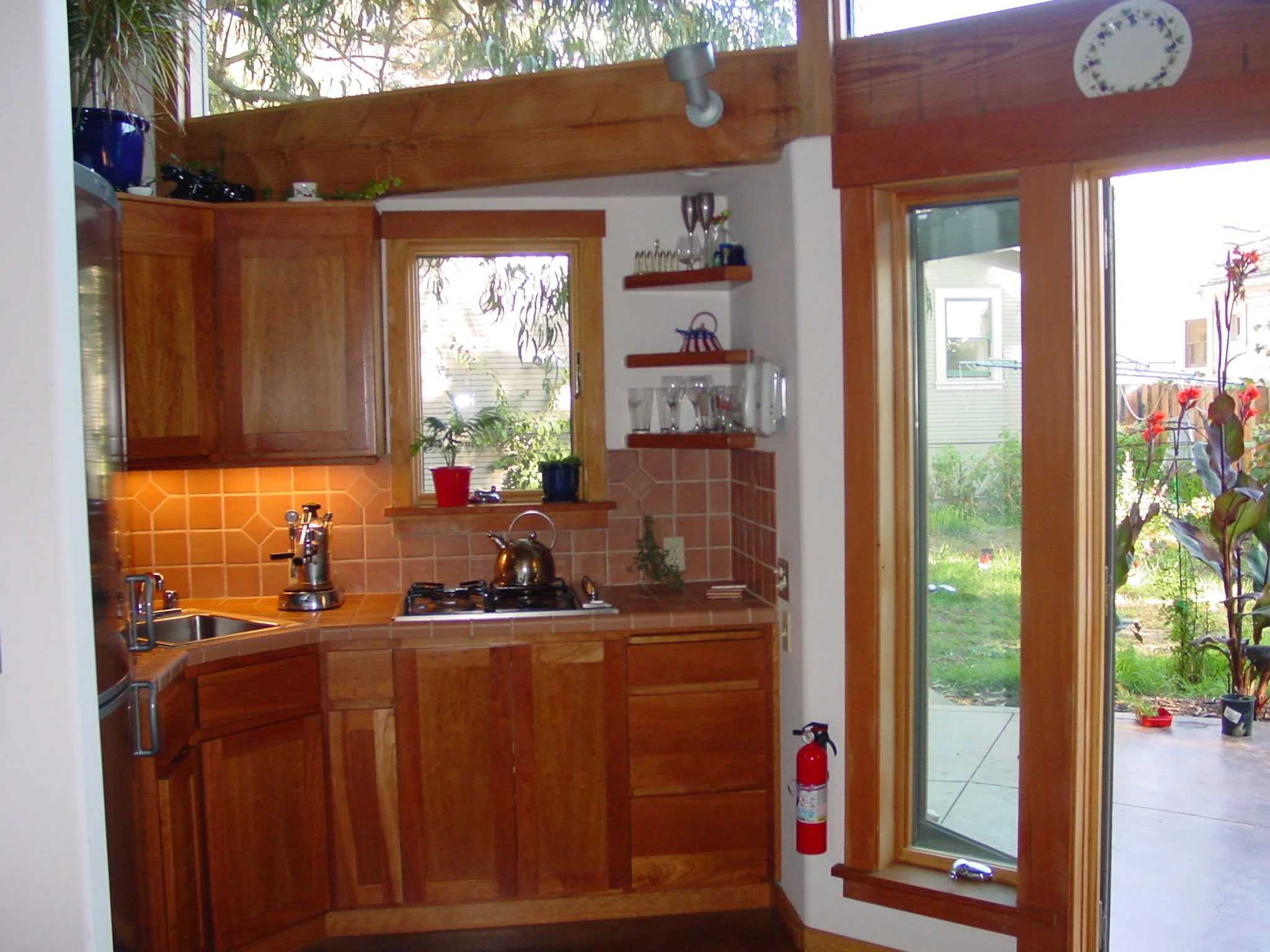 Compact kitchenette with warm wood cabinets, tiled backsplash, and tall windows beside a glass door, showing the small but practical kitchen Levitch designed for the owners cottage.