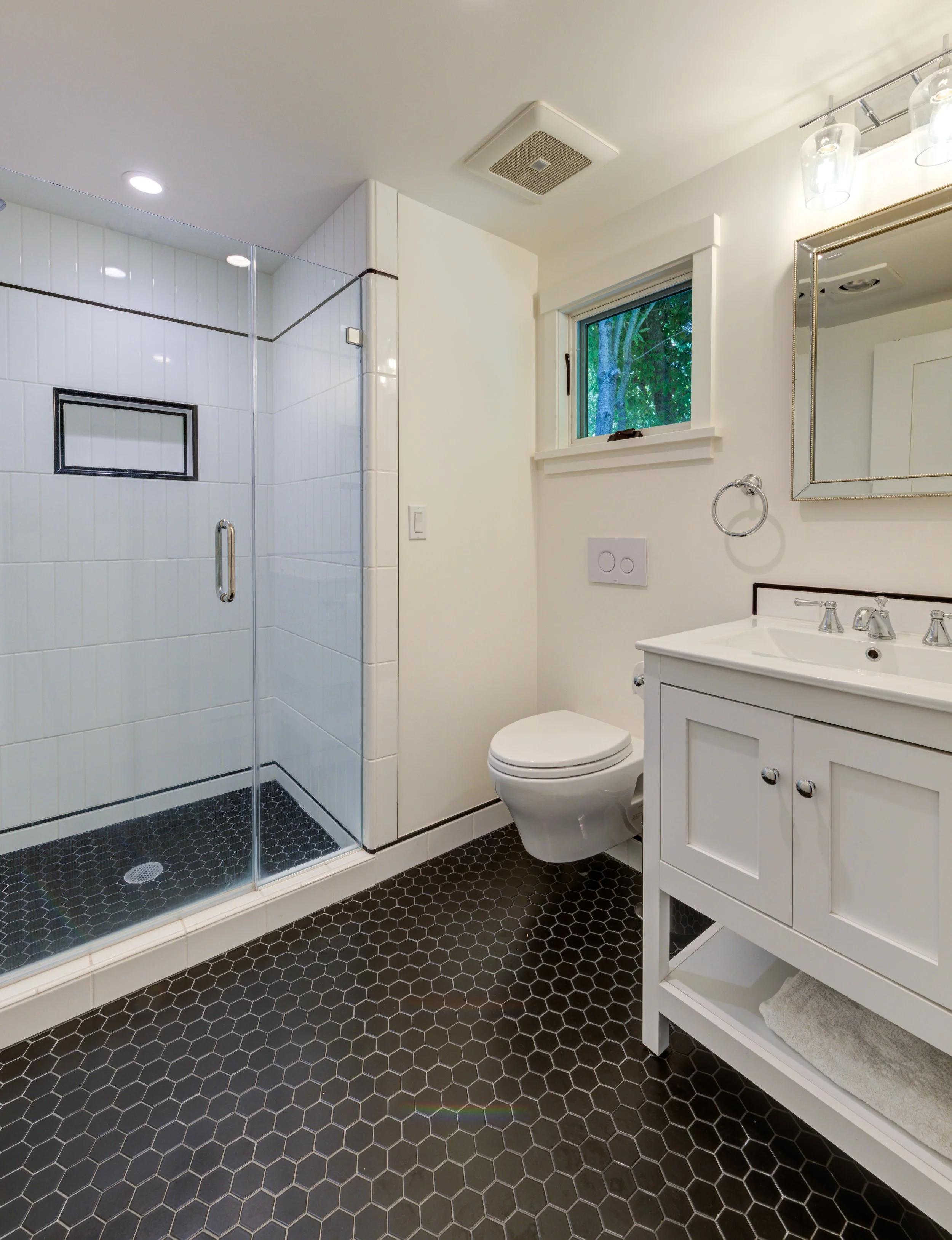 Full Bathroom with white walls, plumbing fixtures and cabinets with a black hex tile floor and accents throughout. Walk-in shower with pivoting glass door.