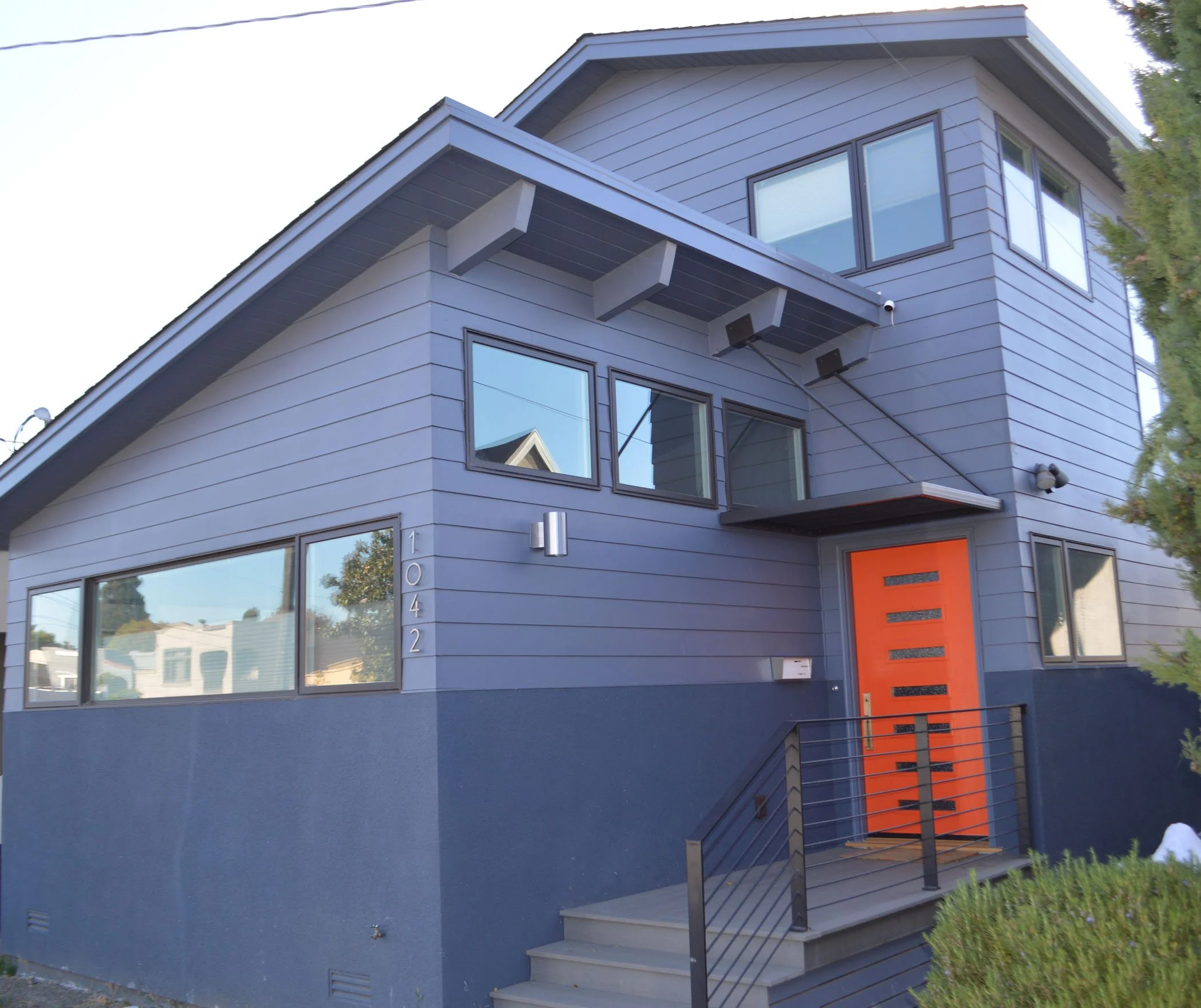 Exterior of the remodeled Albany home with a new second-story addition, blue lap siding, large modern windows, and a bright orange front door with metal entry railing.