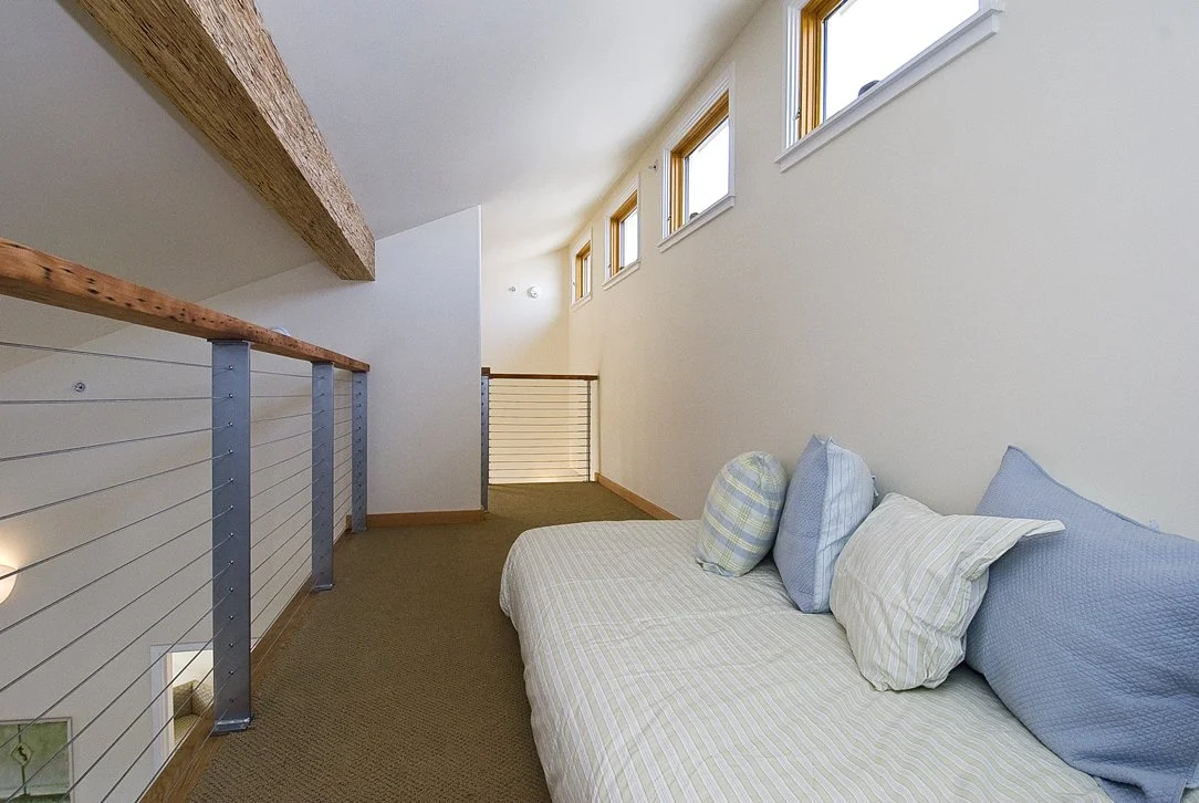 Loft bedroom with built in daybed, exposed wood beams, cable railing, and clerestory windows that bring daylight deep into the space as part of the sustainable home design.