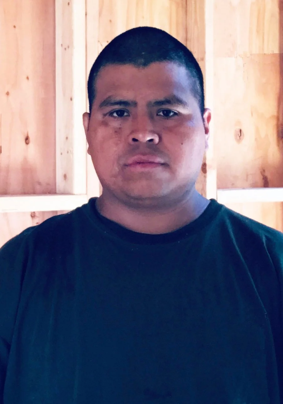 A man with short black hair, wearing a black t-shirt, standing in front of a wooden wall.