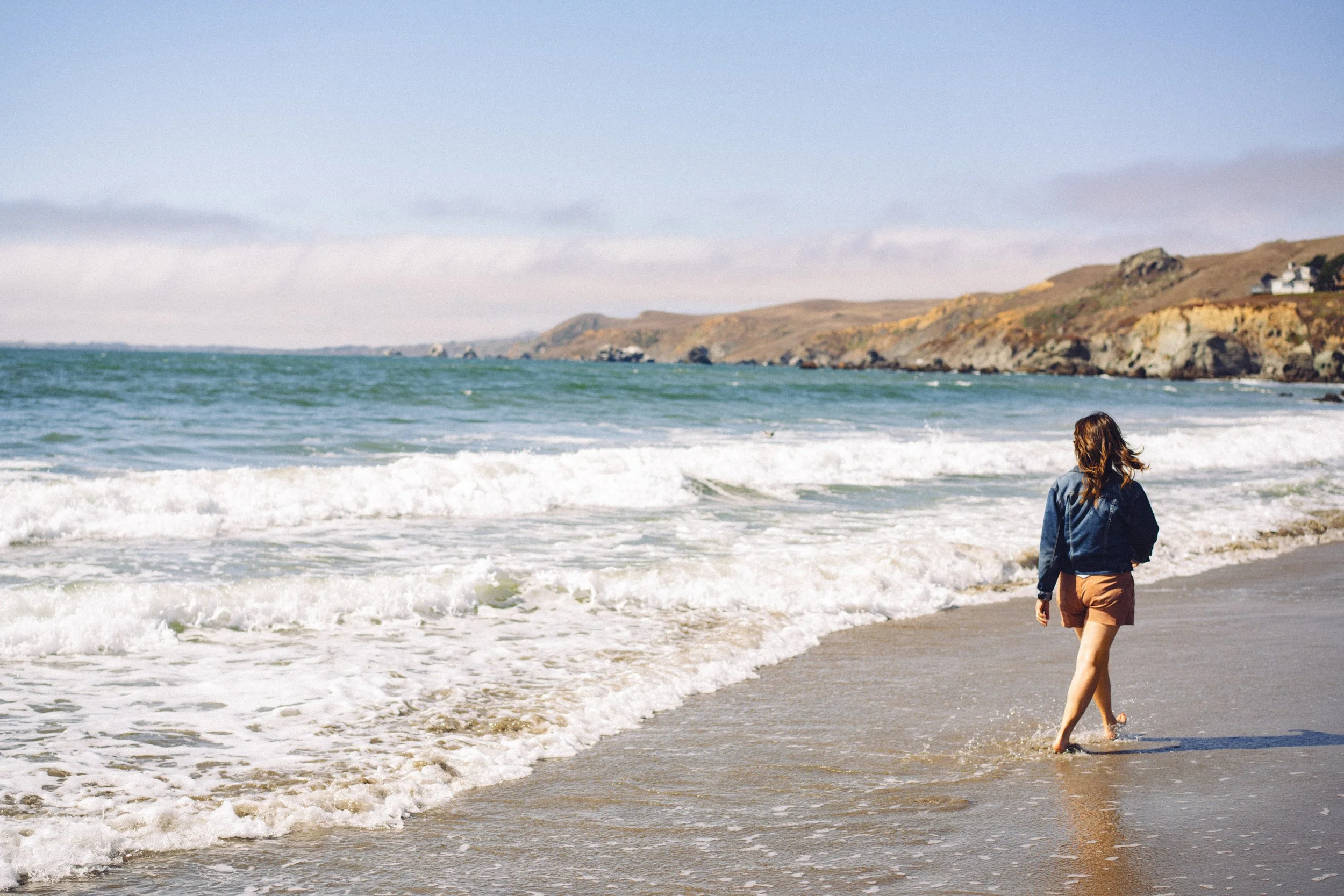 Donna Kato walking along the shoreline at the beach, with waves crashing and a hilly coastline in the distance.