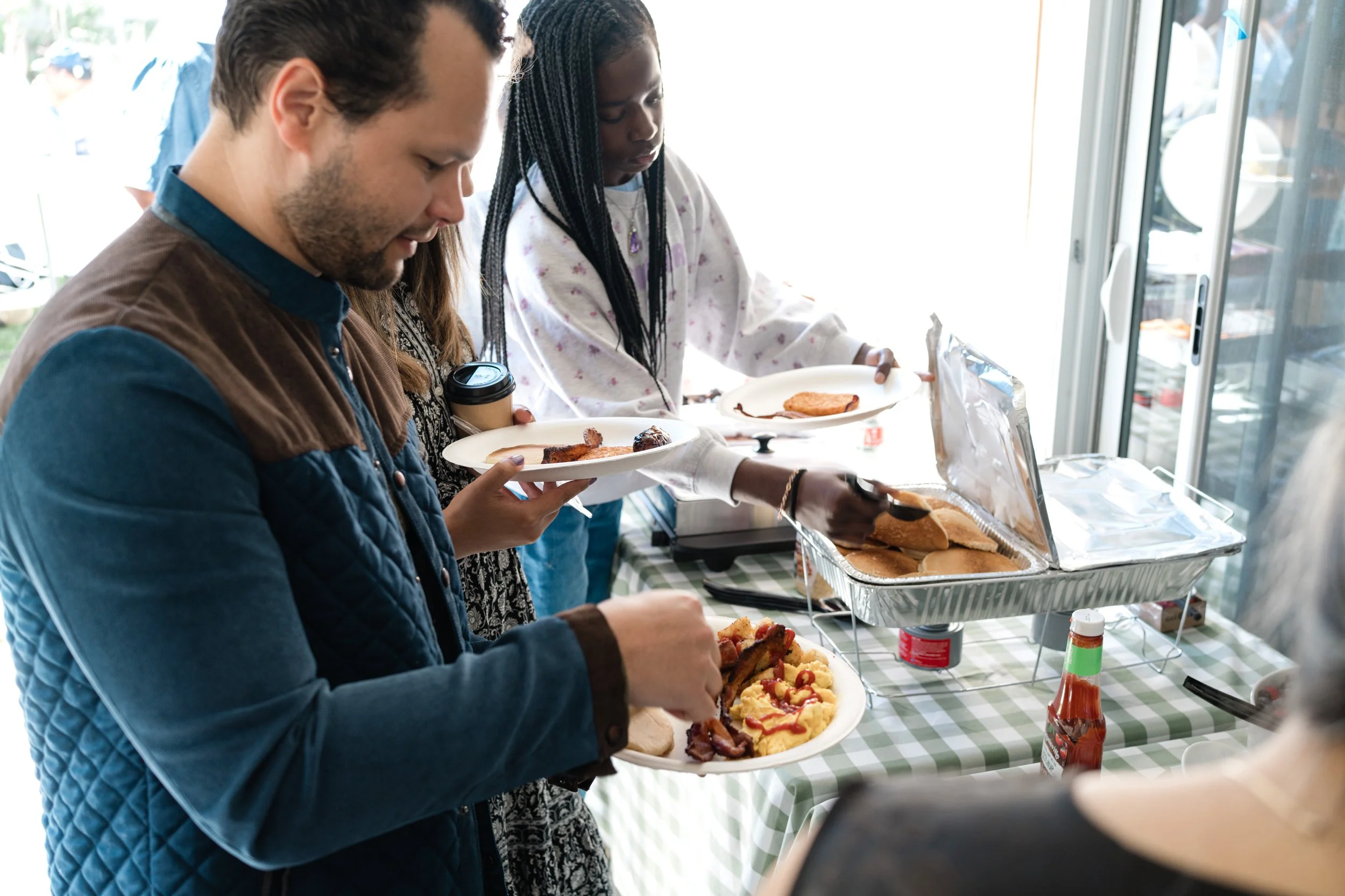 Guests serving themselves from a breakfast buffet with pancakes, scrambled eggs, bacon, and sausage at a pancake-themed first birthday party.