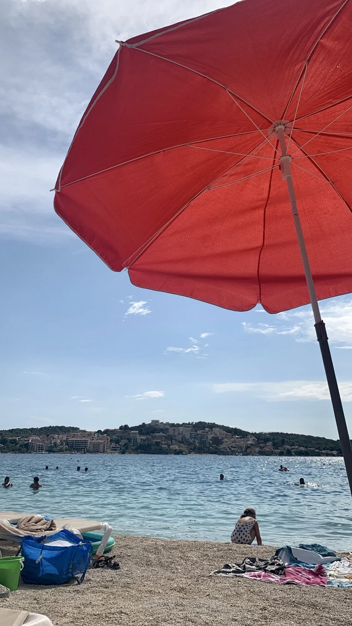 View from beneath a red beach umbrella at a pebble beach in Primošten, Croatia, overlooking the Adriatic Sea and the historic hillside town.
